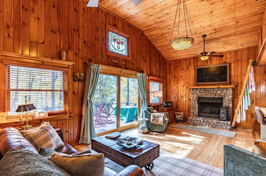 The great room with a stone fireplace, leather sofa, plaid rug, and sliding doors to the deck